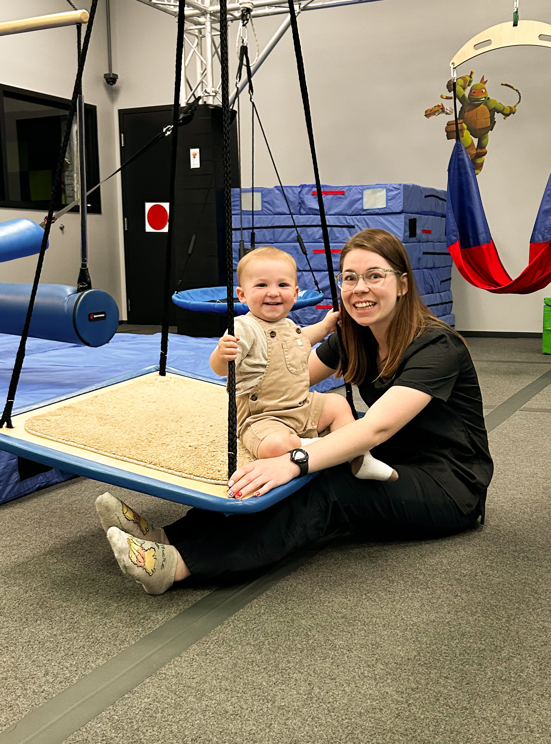 An infant smiling on a platform swing with a therapist
